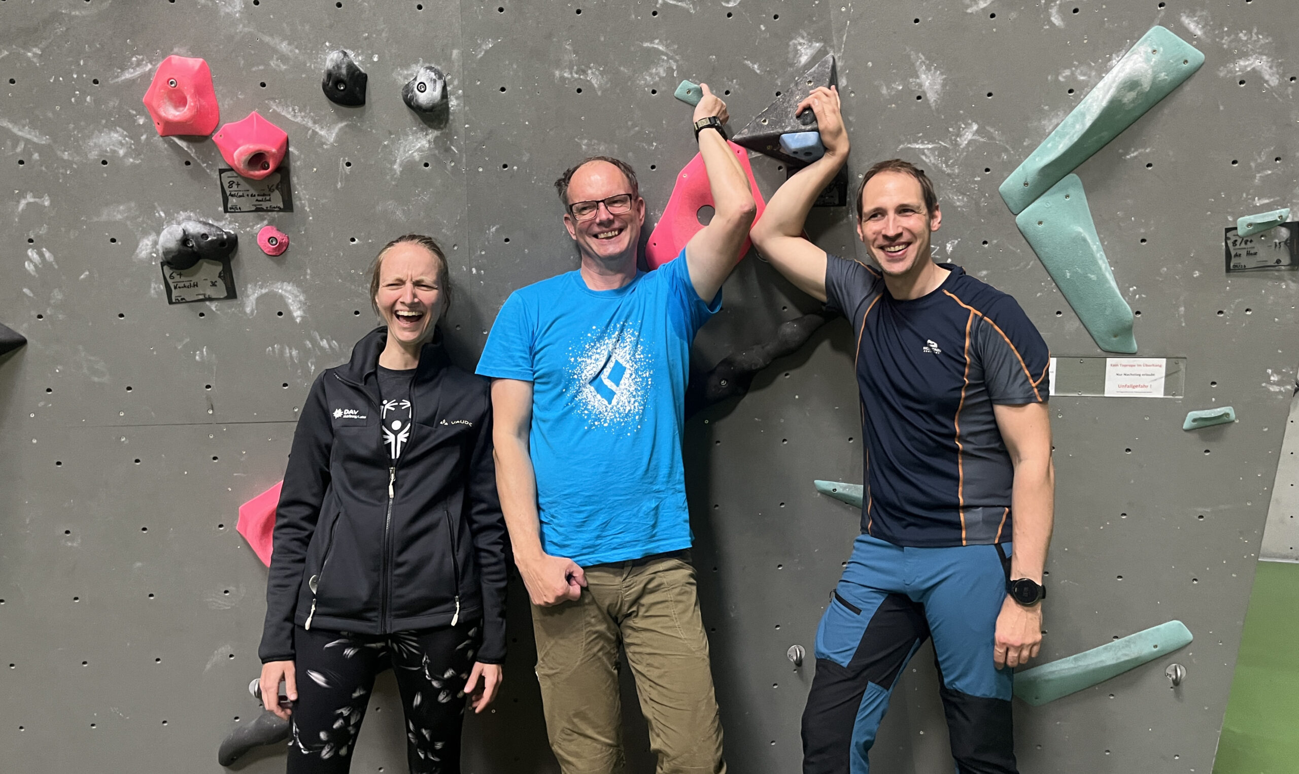 Drei Personen (eine Frau und zwei Männer) stehen vor einer Kletterwand und lachen in die Kamera.