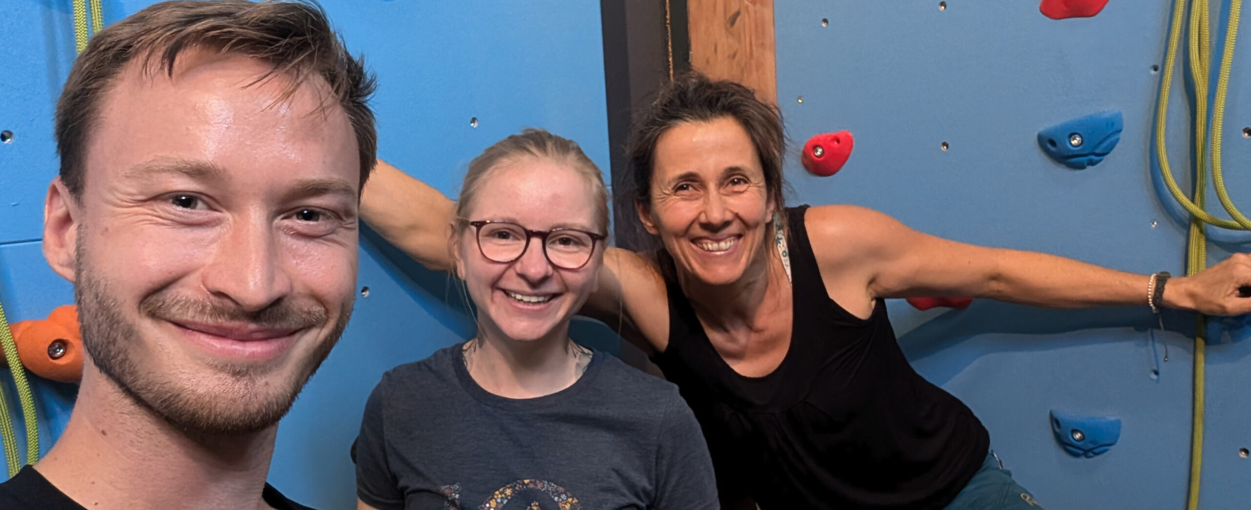 Drei Personen vor der Kletterwand (links ein Mann, rechts zwei Frauen) lachen in die Kamera